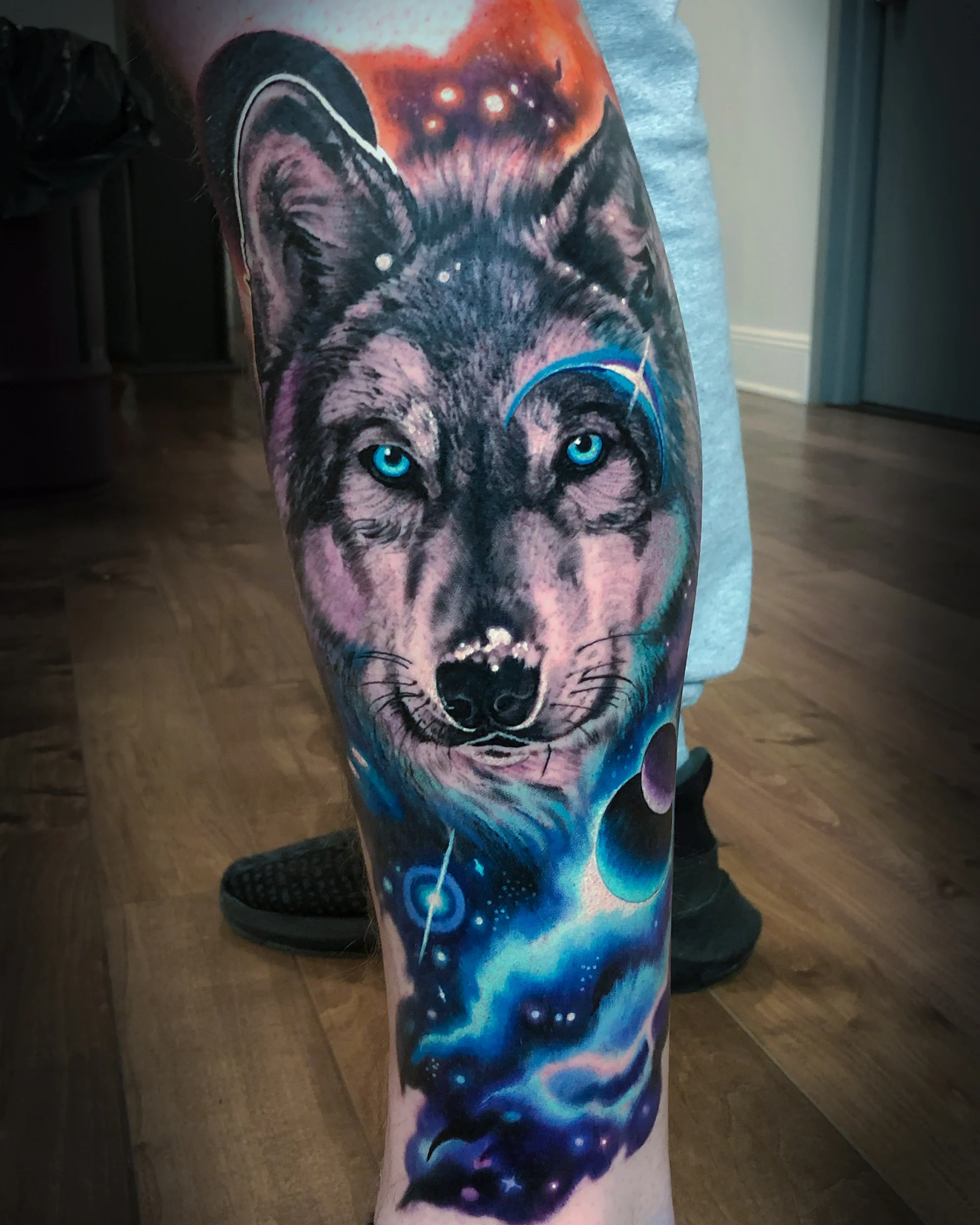 Color realism wolf portrait with glowing blue eyes dissolving into a blue and violet galaxy with planets, calf tattoo by Austin tattoo artist Bobby Cupparo