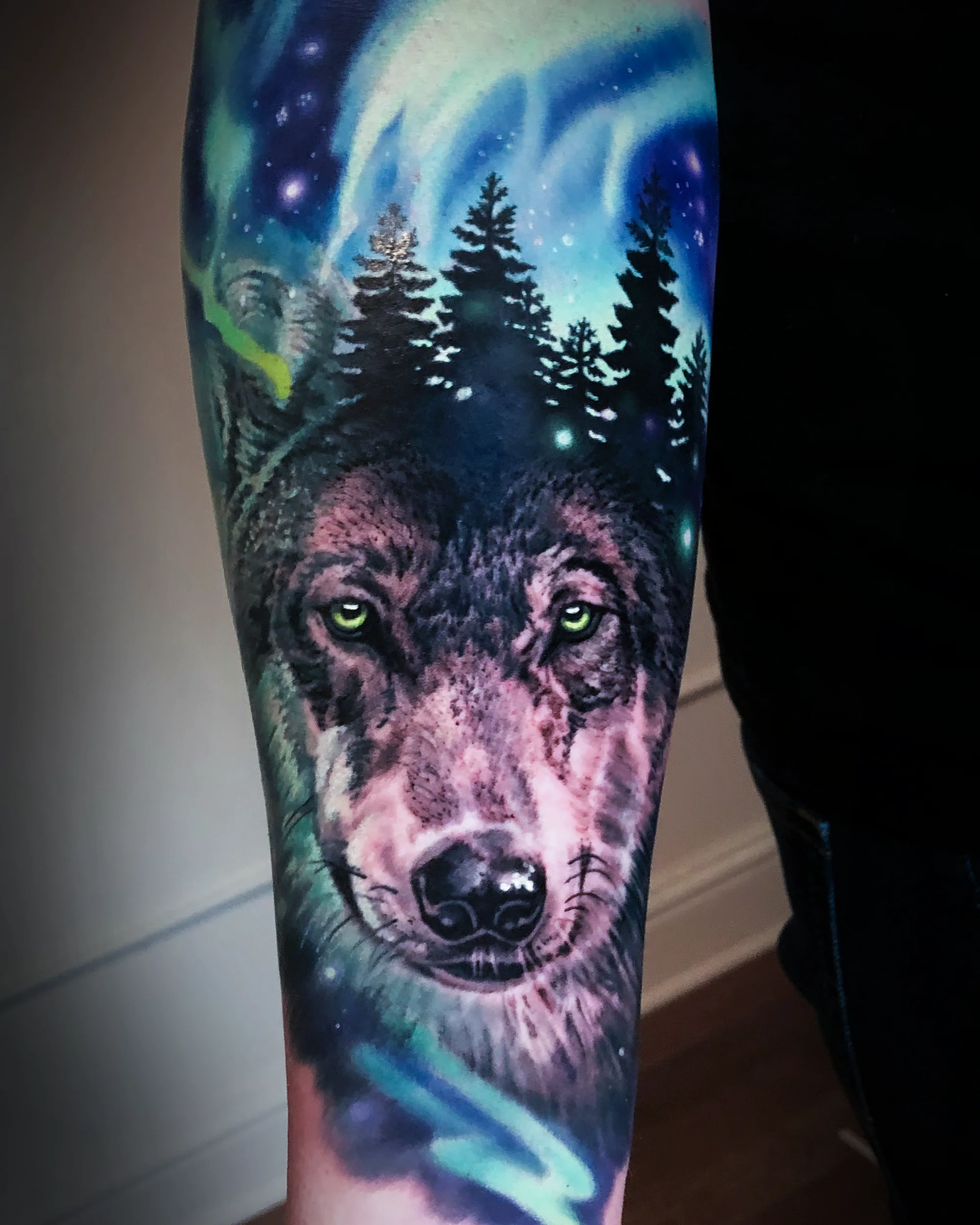Color realism wolf with aurora borealis and pine forest forearm tattoo by Austin tattoo artist Bobby Cupparo